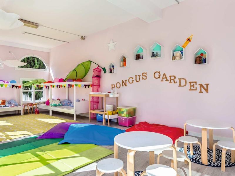 Bunter, heller Kinderraum mit Spielmatten, Tischen, Kissen und Wanddeko "Pongus Garden" an der Wand.
