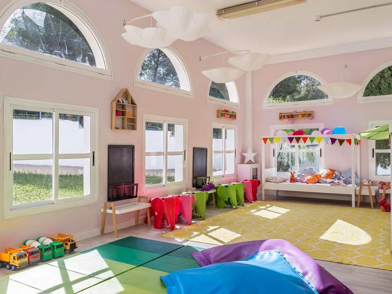 Heller, bunter Kindergartenraum mit großen Fenstern, Teppichen, Spielzeugen und Tischen.