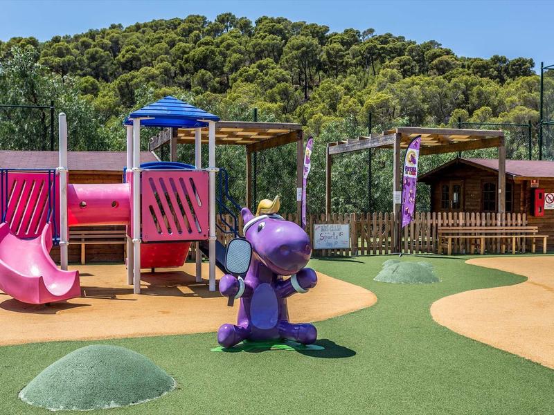 Kinderspielplatz mit Rutschen, einer lilafarbenen Elefantenwippe und Holzhäusern im Hintergrund.