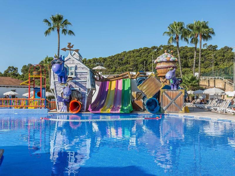 Bunter Wasserspielplatz mit Rutschen und Palmen neben großem blauen Pool unter klarem Himmel.