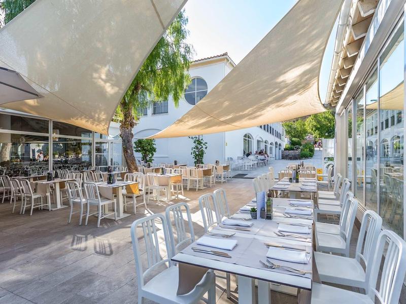 Freiluftrestaurant mit weißen Stühlen und Tischen unter hellen Sonnensegeln am Straßenrand.