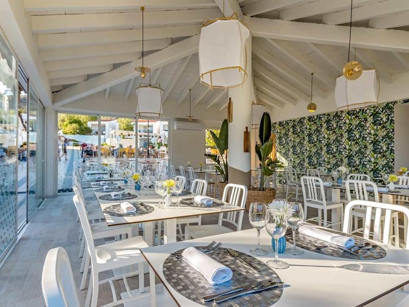 Helles Restaurant mit weißen Möbeln, Holzdecke, Pflanzen und großen Fenstern zum Außenbereich.