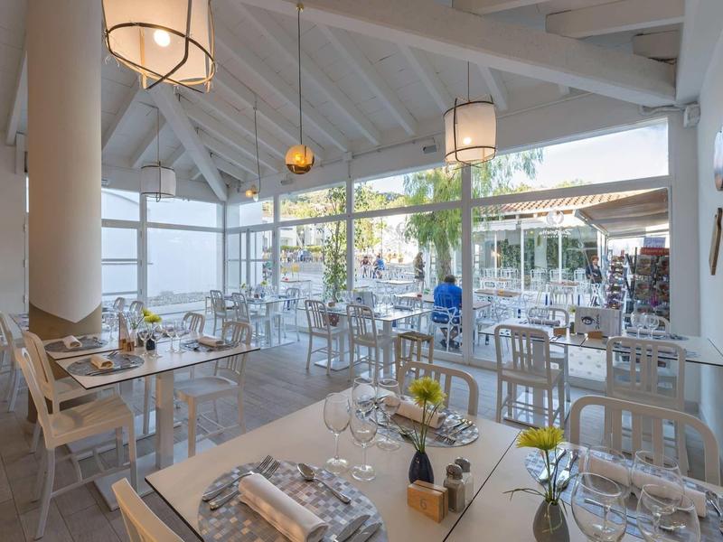 Modernes, helles Restaurant mit großen Fenstern, weißen Möbeln und gedeckten Tischen.