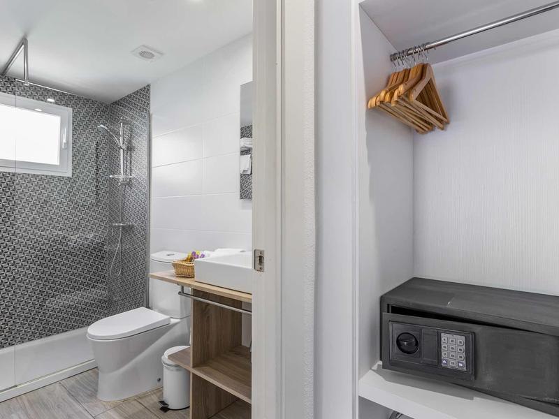 Moderner Badbereich mit Dusche, WC und Waschbecken; daneben ein offener Schrank mit Safe und Kleiderbügeln.