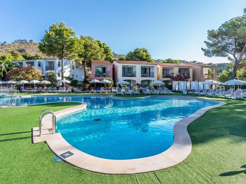 Großer Pool mit klar blauem Wasser, umgeben von grüner Wiese und weißen Gebäuden bei sonnigem Himmel.