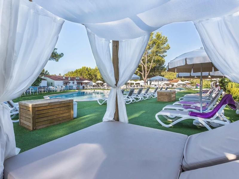 Blick von einem weißen Pavillon mit Vorhängen auf grüne Liegewiese und Sonnenliegen.
