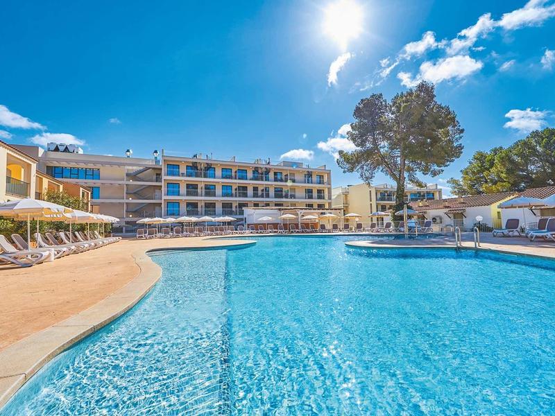 Großer blauer Pool vor modernem Hotelgebäude mit Liegestühlen und Bäumen bei sonnigem Himmel.