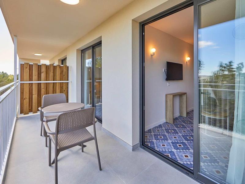 Großer Balkon mit grauem Boden, zwei Stühlen, Glastür und Blick auf Bäume und Sonnenlicht.