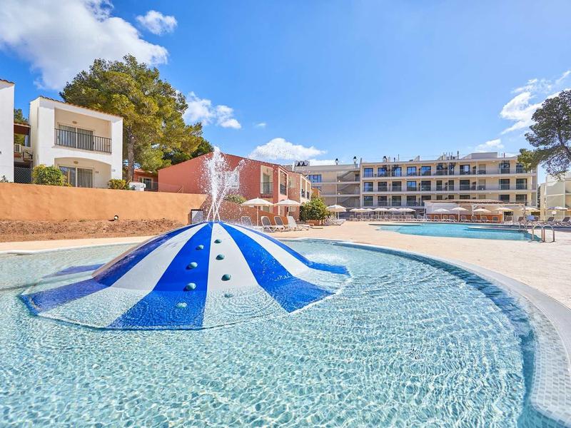 Kinderbecken mit blau-weißem Wasserspiel vor großen weißen Ferienhäusern unter blauem Himmel.