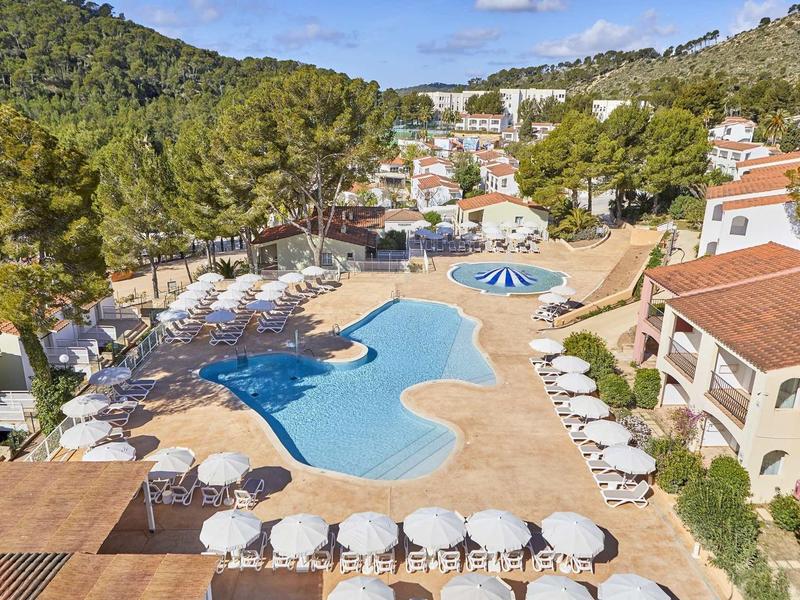 Großer Pool mit Sonnenschirmen und Liegen, umgeben von Häusern und grünen Hügeln unter blauem Himmel.