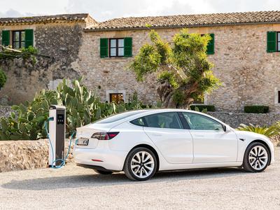 Weißes Elektroauto lädt an Ladestation vor rustikalem Steinhaus mit grünem Fensterladen.