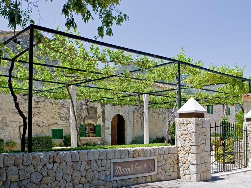 Kamienny wejście do hotelu z zielonym listowiem i stelażem na daszki przeciwsłoneczne.