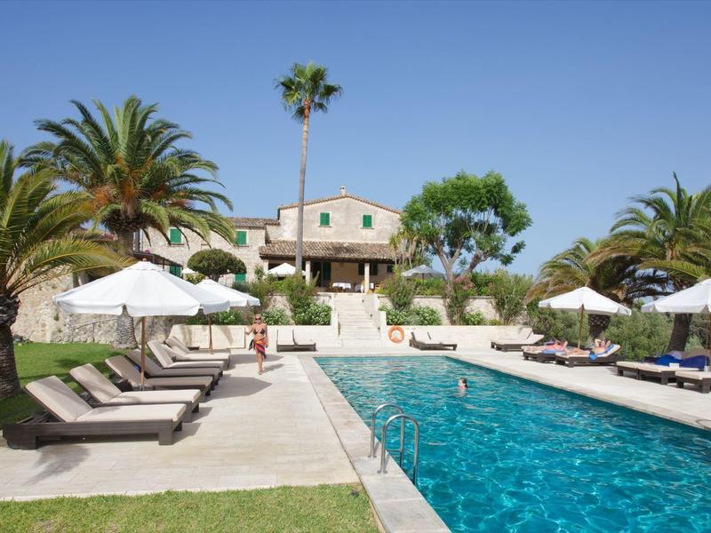 Duży basen z leżakami i palmami przed dwupiętrowym domem wakacyjnym.