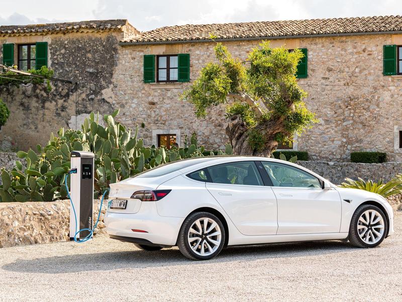 Biały samochód elektryczny ładuje się na stacji przed rustykalnym kamiennym domem z zielonymi okiennicami.