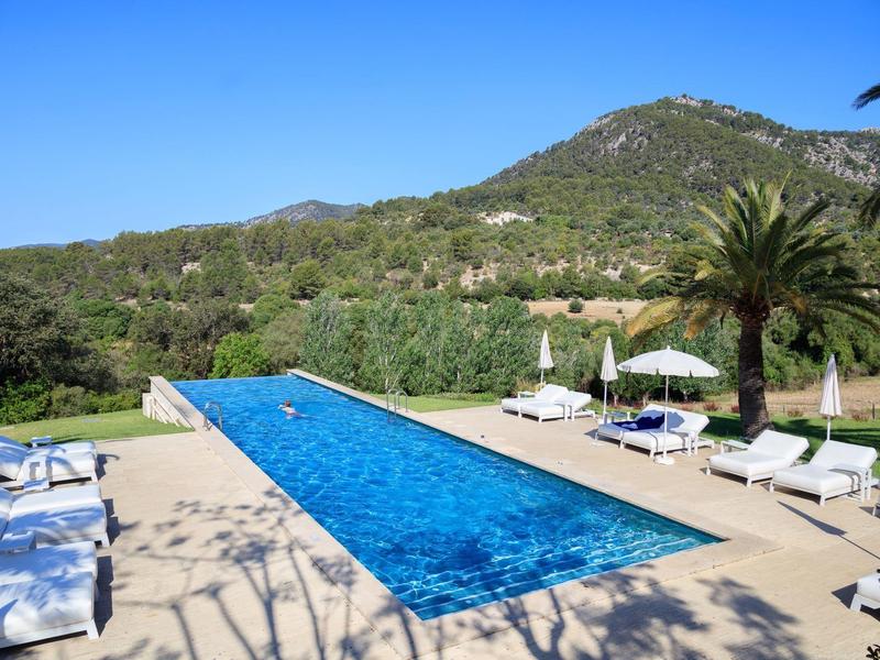 Długi, wąski basen z leżakami i parasolami na tle zalesionych wzgórz pod czystym niebem.