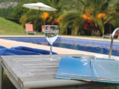 Verre de vin et livre ouvert sur une table en bois près de la piscine avec parasol et palmiers.