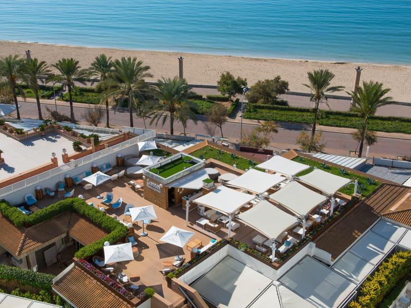 Vogelperspektive auf eine moderne Strandbar mit Sonnenschirmen, Palmen und Blick auf das blaue Meer.