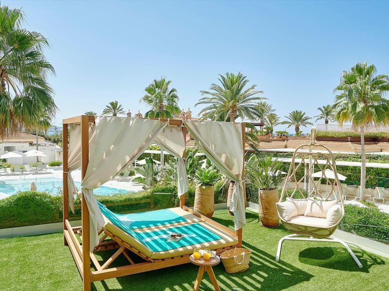 Grüne Rasenfläche mit weißem Baldachinbett, Schaukelstuhl und Palmen im sonnigen Garten.