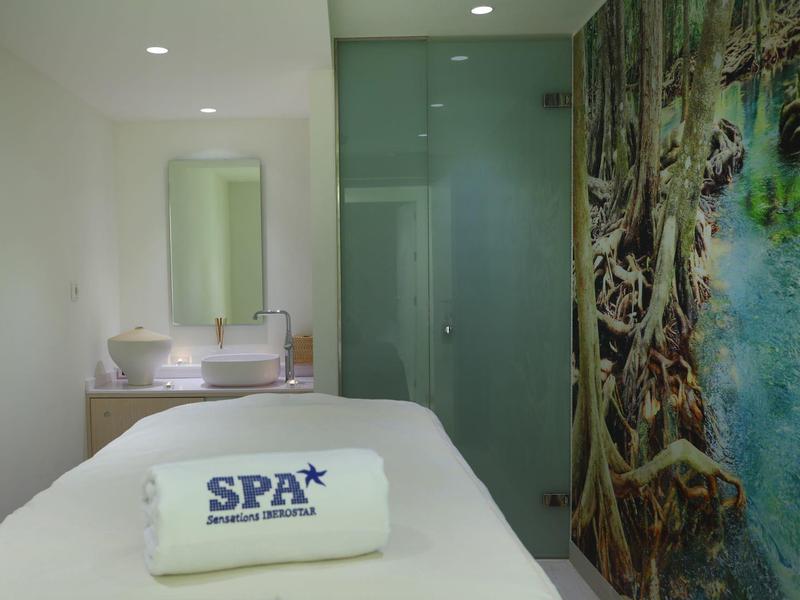 Weißes Spa-Zimmer mit Liege, Handtuch, Spiegel und Wandbild eines grünen Waldes am rechten Rand.