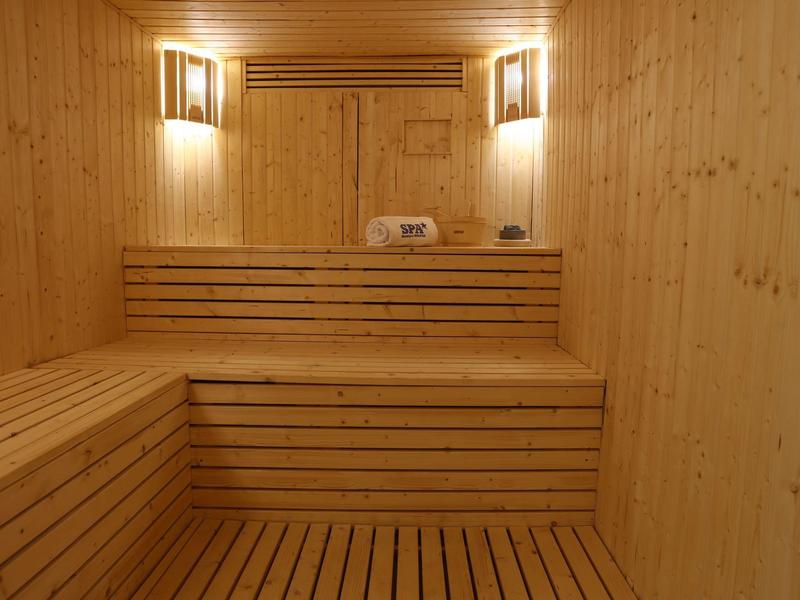 Holzsauna mit zwei Sitzebenen, Wandleuchten und Saunaofen, helle Holzvertäfelung.