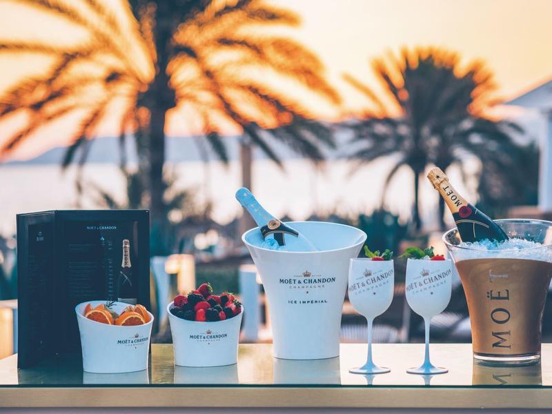 Bar mit Eiskübeln, Cocktails und Früchten vor Palmen und Sonnenuntergang am Strand.
