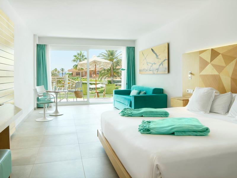 Helles Hotelzimmer mit Doppelbett, grünem Sofa, Tisch, großem Fenster und Blick auf Garten und Pool.