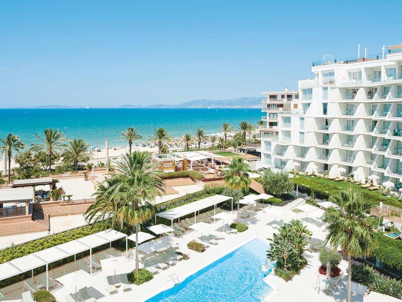 Hotel mit großem Pool, zahlreichen Liegestühlen und Palmen am Meer bei klarem Himmel.