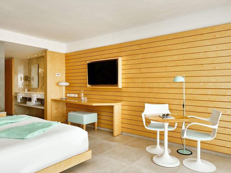 Helles Hotelzimmer mit Holzwand, großem Bett, Fernseher, Schreibtisch und zwei weißen Stühlen mit kleinem Tisch.