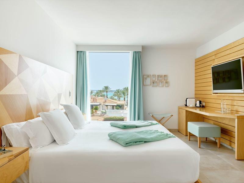 Helles Hotelzimmer mit Doppelbett, Holzwand, Schreibtisch, Fernseher und Balkon mit Meerblick.