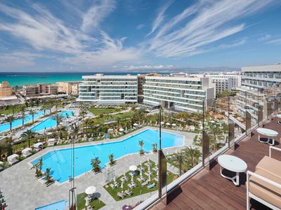 Complesso alberghiero moderno con più piscine e vista sul mare sotto un cielo azzurro.