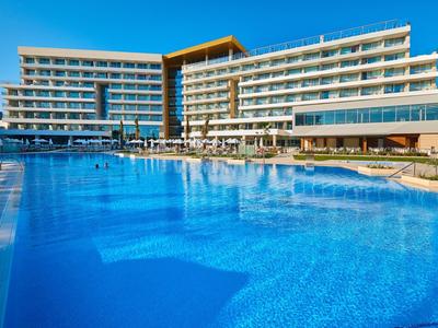 Modernes Hotel mit großem, klarem Außenpool und mehreren Etagen in hellem Beige bei blauem Himmel.
