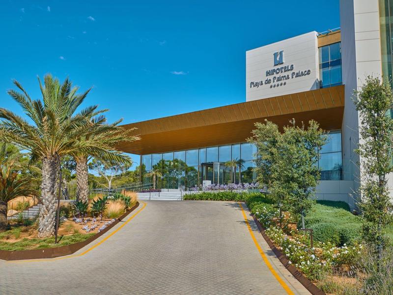 Ingresso moderno dell'hotel con viale coperto e palme sotto un cielo azzurro.