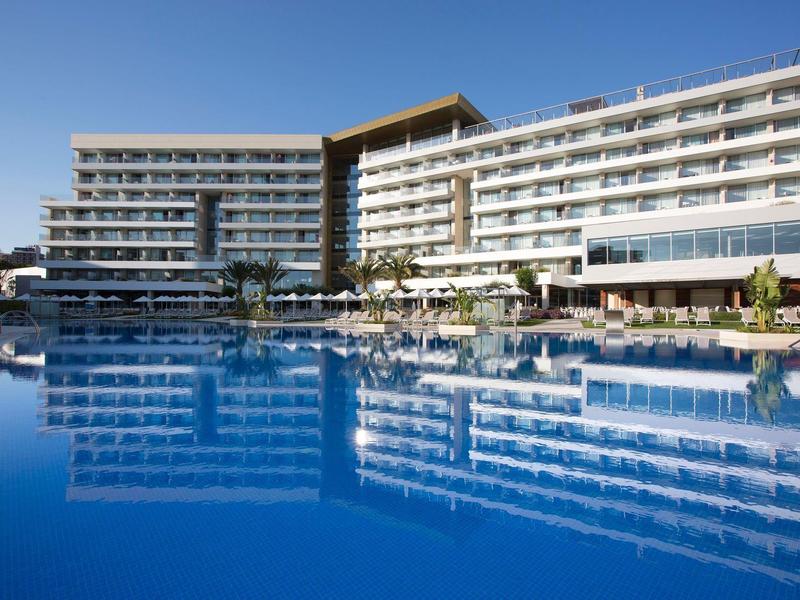 Grande hotel su più piani accanto a una piscina blu sotto un cielo azzurro.