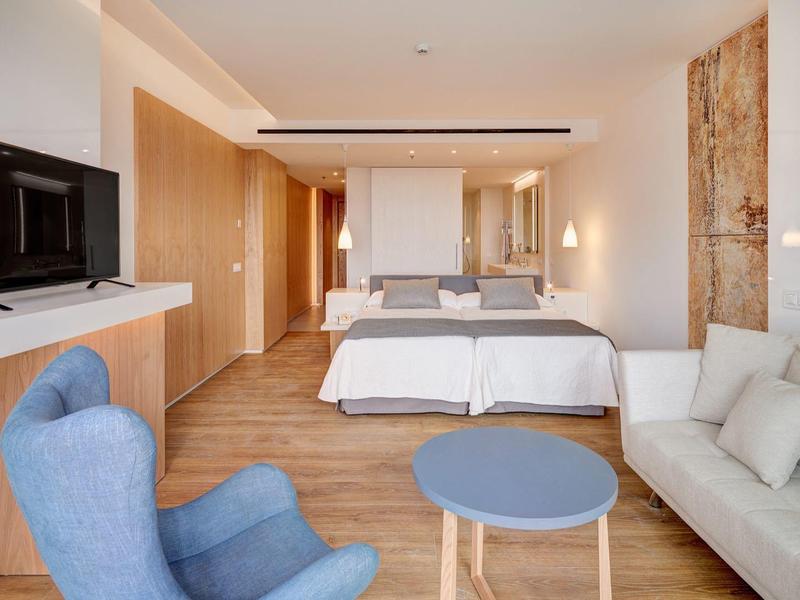 Ampia camera d'hotel con letto matrimoniale, divano, poltrona blu, tavolo rotondo e TV.