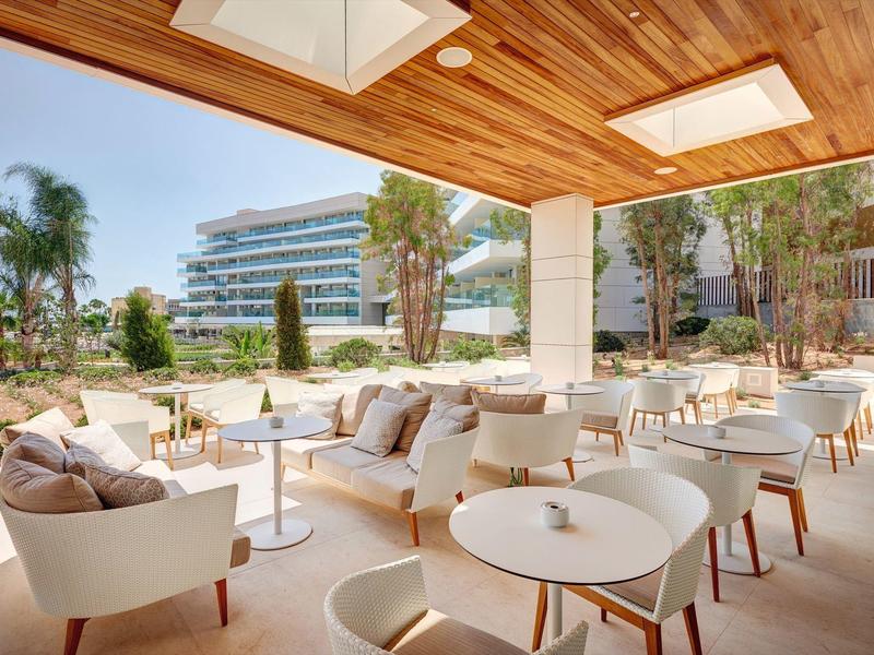 Area salotto aperta con sedie moderne e divani sotto una tettoia in legno, circondata da palme e edifici.