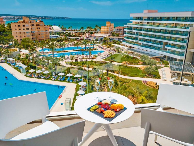 Vista da un balcone su un resort con piscine, giardino e mare sullo sfondo.