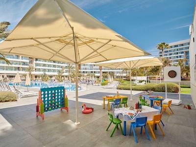 Outdoor-Spielbereich mit bunten Kindertischen, Stühlen und Spielen neben Hotelpool unter Sonnenschirmen.