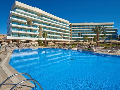 Moderner Hotelkomplex mit blauem Pool, Liegestühlen und Palmen bei klarem Himmel.