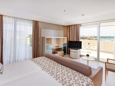 Helles Schlafzimmer mit großem Bett, Tisch, Stuhl, Fernseher und bodentiefen Fenstern mit Blick.