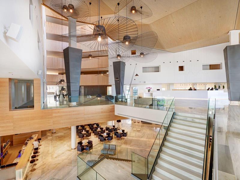 Ingresso di hotel moderno e luminoso con pannelli in legno, scala e aree salotto al piano inferiore.