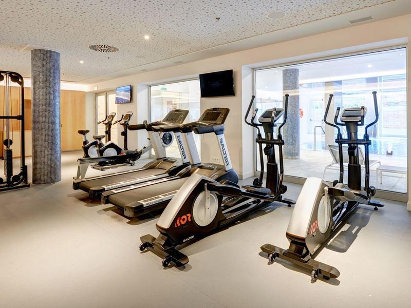 Palestra moderna con tapis roulant e cyclette ellittiche, pareti chiare e grandi finestre.