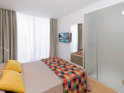 Modern hotel room with double bed, colorful blanket, yellow pillows, frosted glass door, and window curtains.