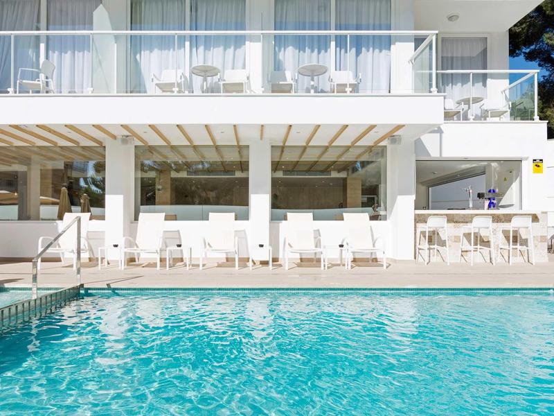 Clear blue pool in front of a modern white hotel with outdoor seating and glass balcony.