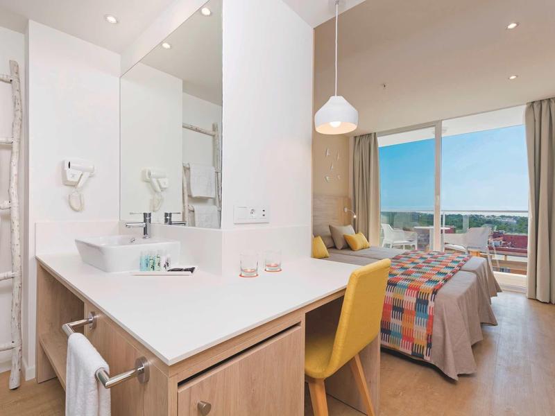 Bright hotel room with desk, yellow chair, bed, and large window with city view.
