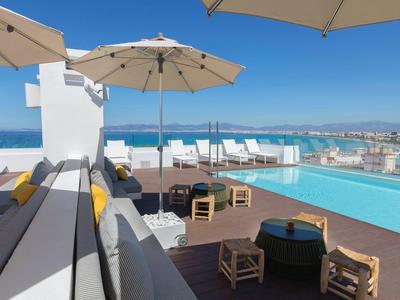 Taras na dachu z basenem, leżakami i parasolami z widokiem na miasto i morze.