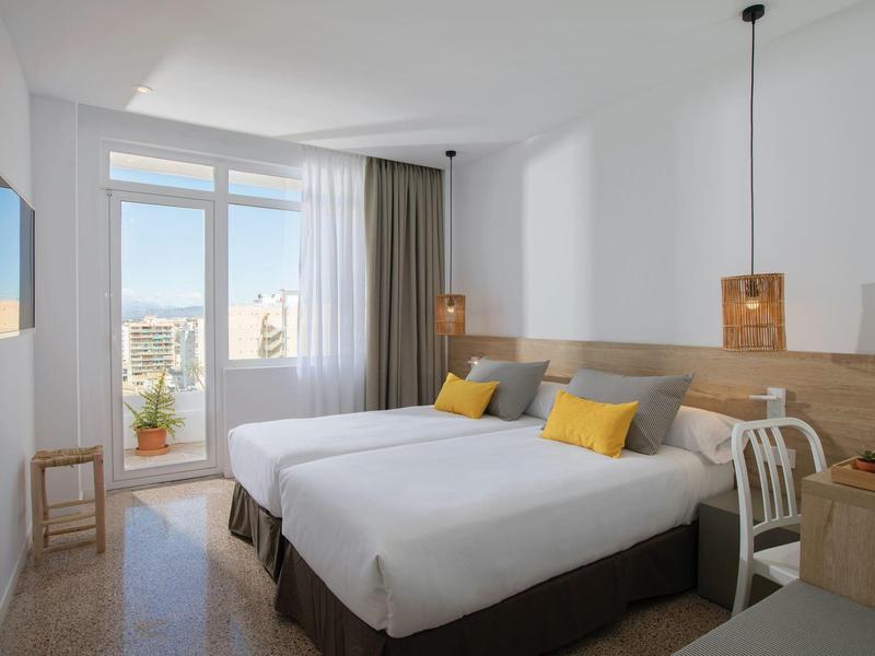 Jasny pokój hotelowy z dwoma łóżkami, szarymi zasłonami, żółtymi poduszkami i widokiem na miasto.