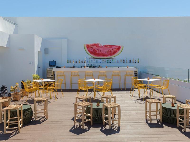 Taras na dachu z barem, drewnianymi stołami i krzesłami, ozdobiony muralem arbuza.