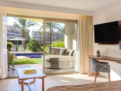 Chambre d'hôtel lumineuse avec vue sur le jardin, coin salon extérieur et piscine visible par de grandes portes coulissantes.