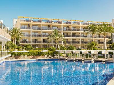 Hôtel à plusieurs étages avec balcons à côté d'une grande piscine avec chaises longues et palmiers.