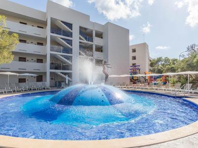 Piscine ronde d'hôtel avec jeux d'eau et chaises longues autour, bâtiment à plusieurs étages en arrière-plan